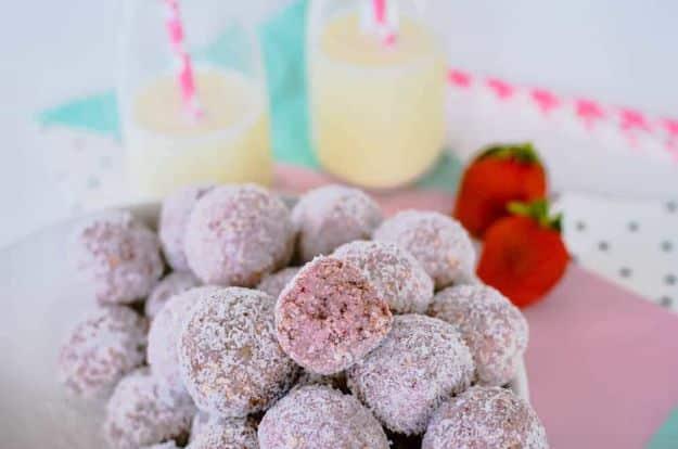 Strawberry bliss balls rolled in coconut piled on a plate, one bitten open, berries and two drinks behind