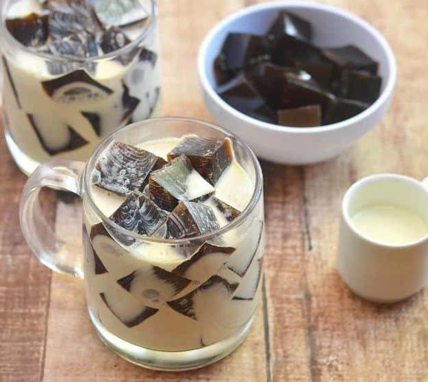 Coffee jelly cubes floating in pale cream in a glass mug, a bowl of dark cubes and a cream pitcher on wood