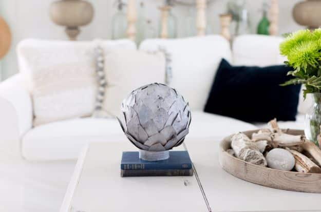 A gray sphere covered in overlapping metal-look leaves rests in a wooden bowl on a neutral styled shelf