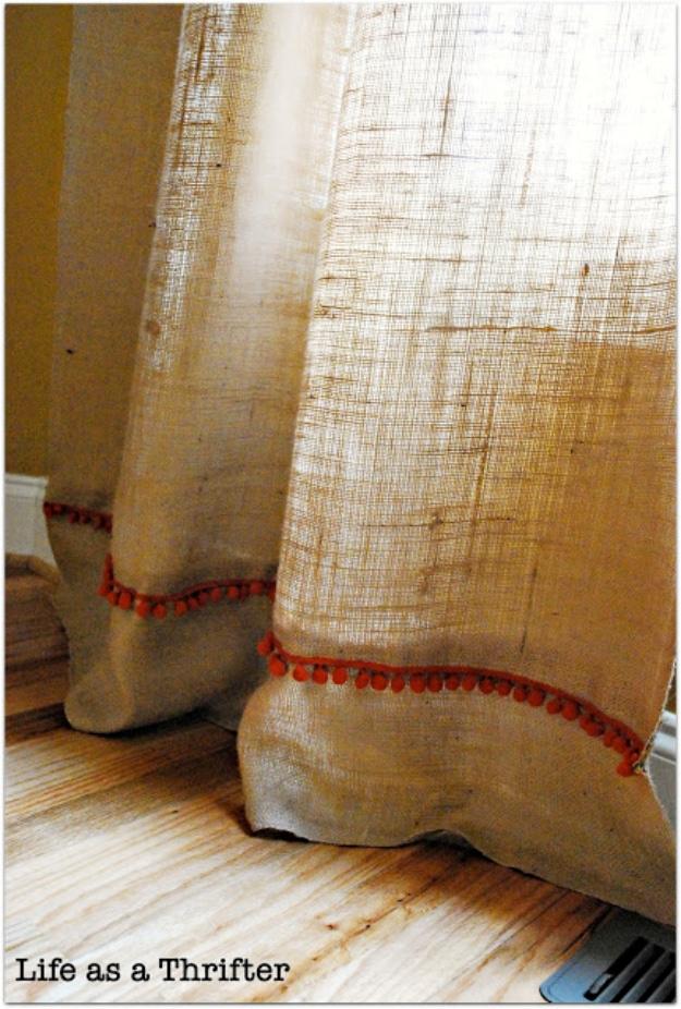 Natural burlap curtain panels puddling on a wood floor, finished with a line of orange pom pom trim along the hem