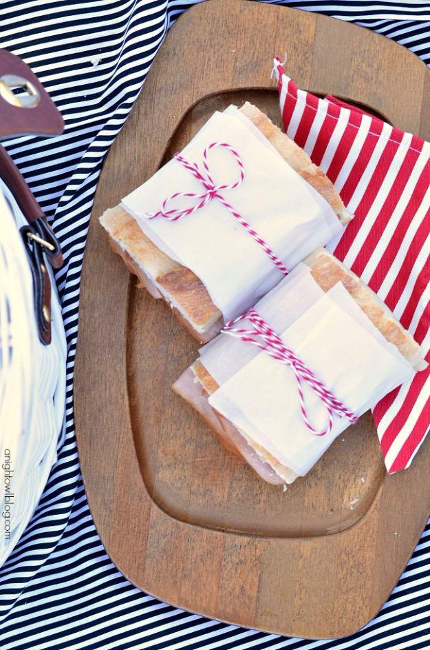 Sandwiches wrapped in parchment and baker's twine for a picnic, tied and stacked on a wooden board over a striped cloth