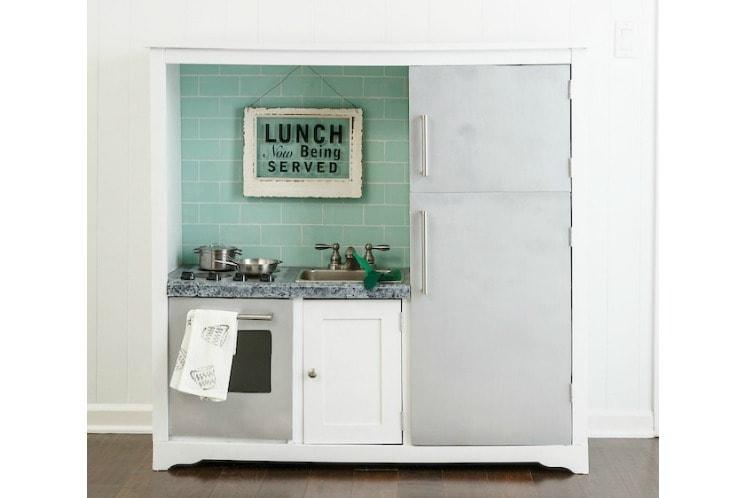 A white play kitchen made from an entertainment center with green tile backsplash, sink, faucet, oven door and silver fridge