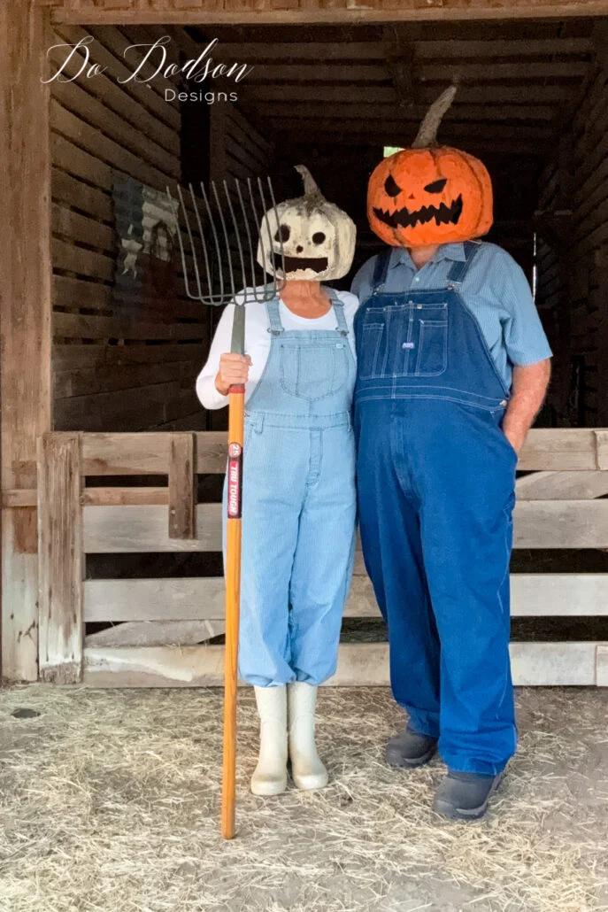 Two adults wearing full costumes with orange and pale paper mache pumpkin heads in front of a rustic barn