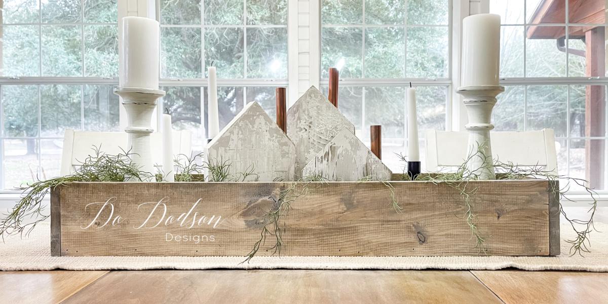 A long stained scrap wood box holding chippy white wooden houses, white taper candles and Spanish moss on a woven runner