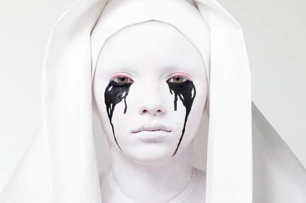 Weeping nun Halloween makeup with white face paint, black tear streaks, red rimmed eyes and a white hood