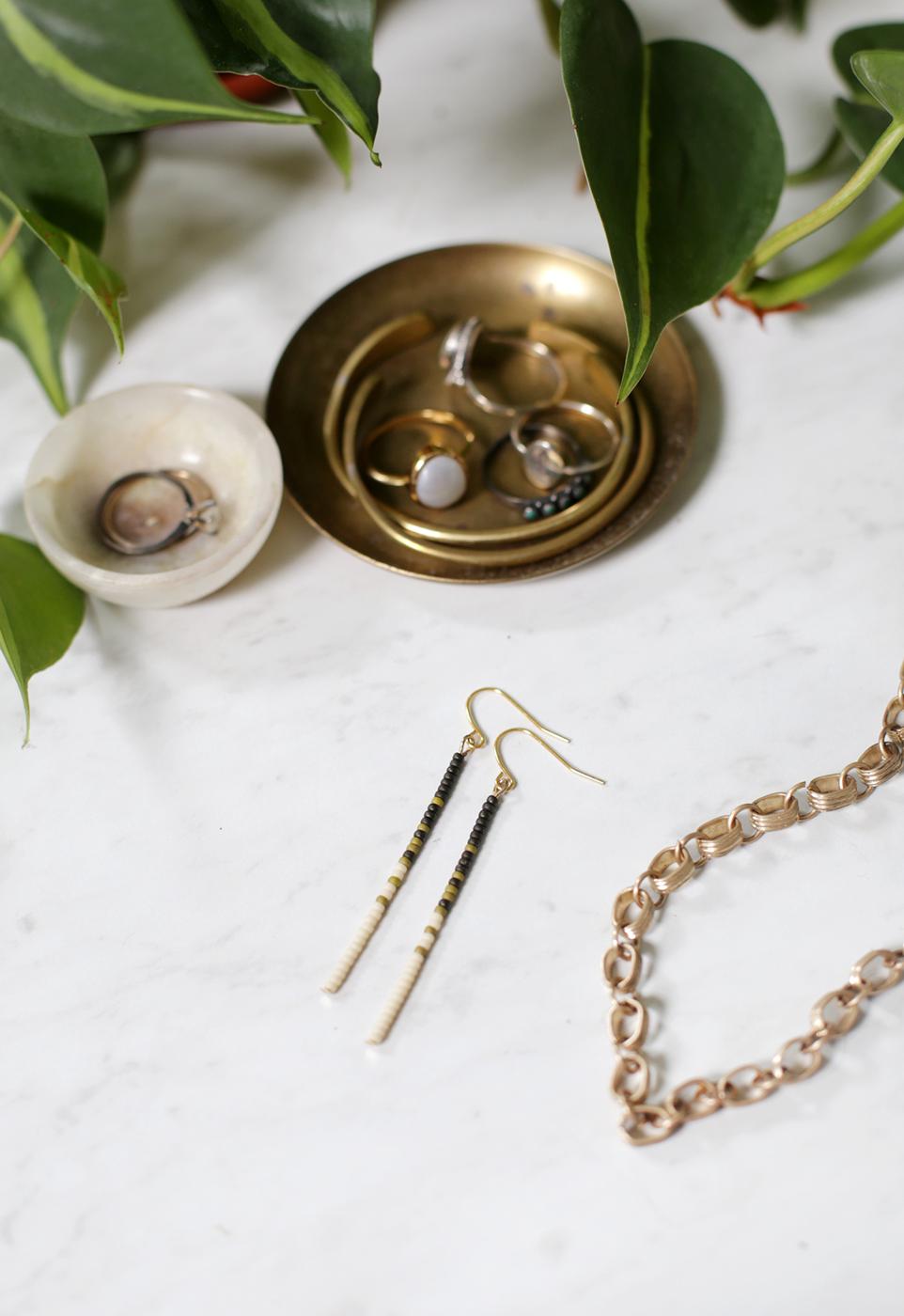 Pair of slim gold and black beaded drop earrings laid beside a brass chain and a small brass dish with a plant leaf