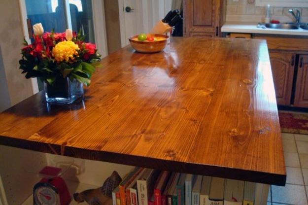 Glossy stained wood countertop on a kitchen island with a knife block, a bowl of fruit and a vase of red and yellow flowers