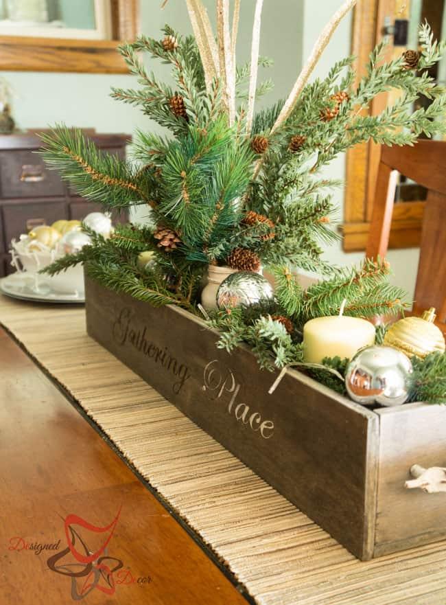 A stained wood box stenciled Gathering Place filled with faux pine, pinecones, silver and gold ornaments and a cream candle