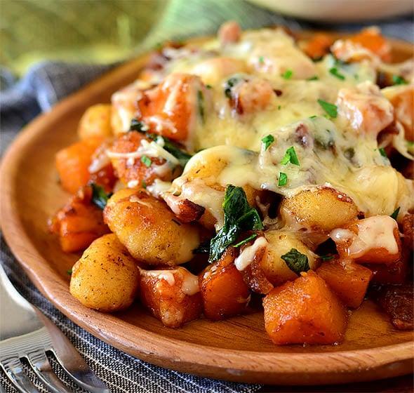 Butternut squash gnocchi with bacon and greens under melted cheese, served on a round wooden plate with a fork