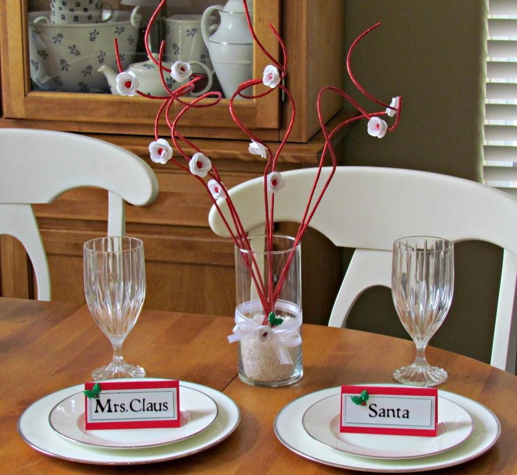 Red glitter branches with white paper flowers in a glass vase of snow between place cards for Santa and Mrs. Claus