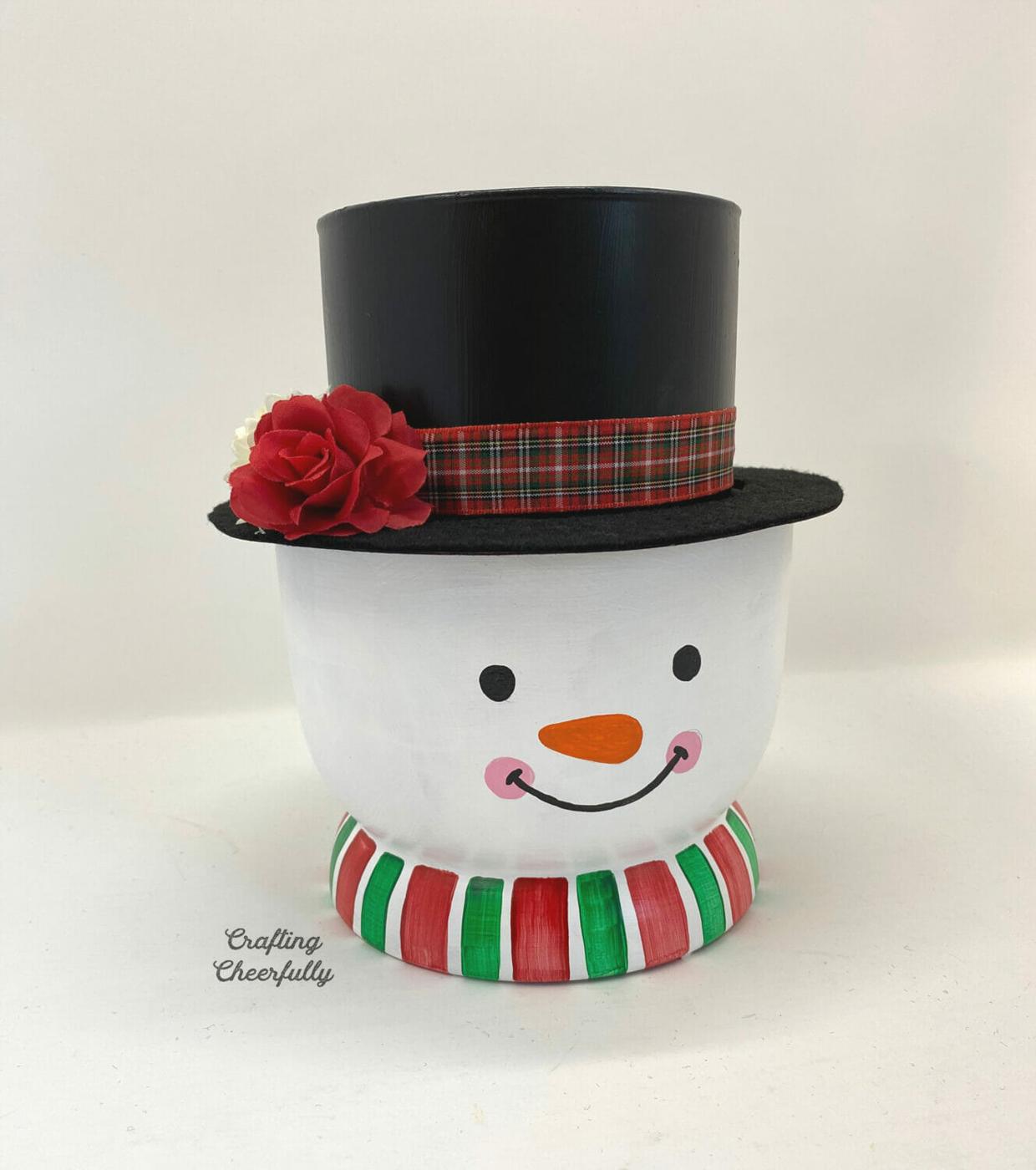 Terracotta pot painted as a snowman face with a black vase top hat, red plaid ribbon and evergreen stems in the hat