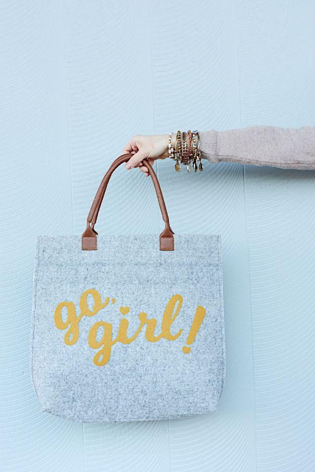 Pale gray felt tote bag lettered go girl in gold with slim leather handles, hanging against a light blue wall