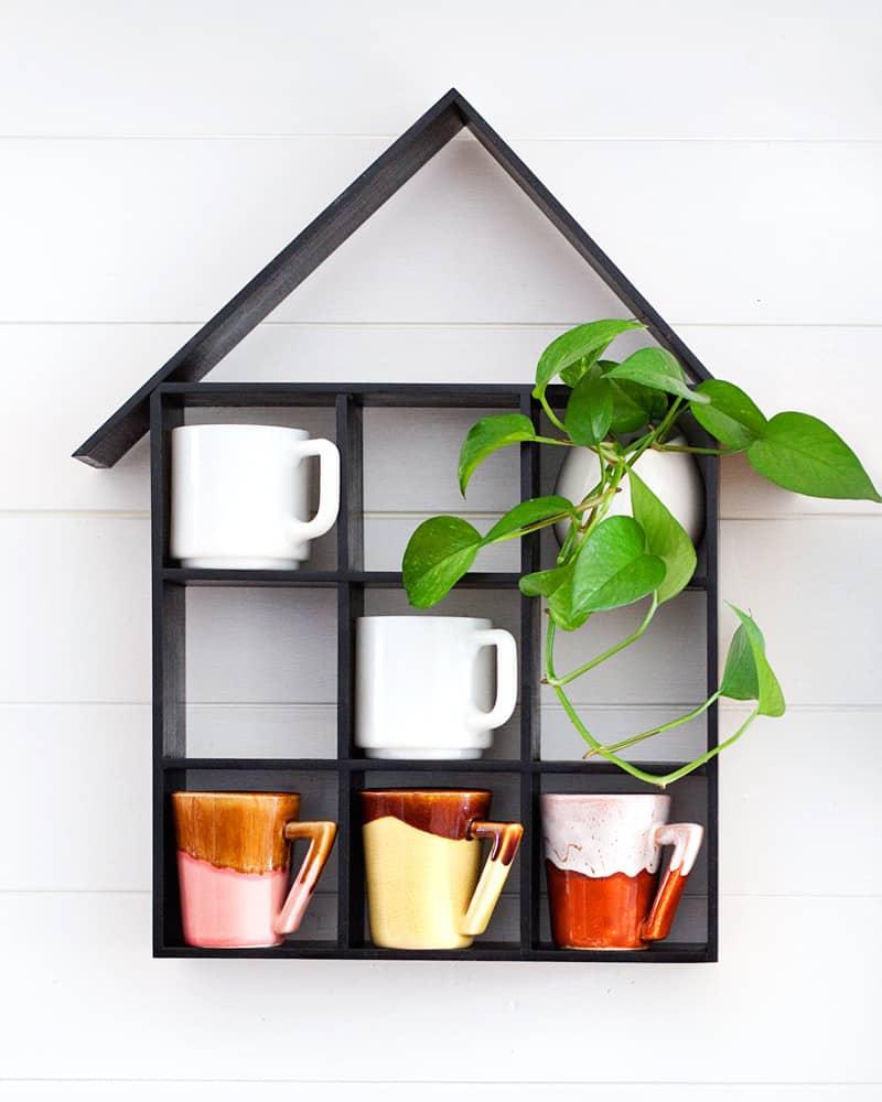 Black house shaped wood shelf with a grid of small square cubbies holding tiny plants and trinkets on a white wall
