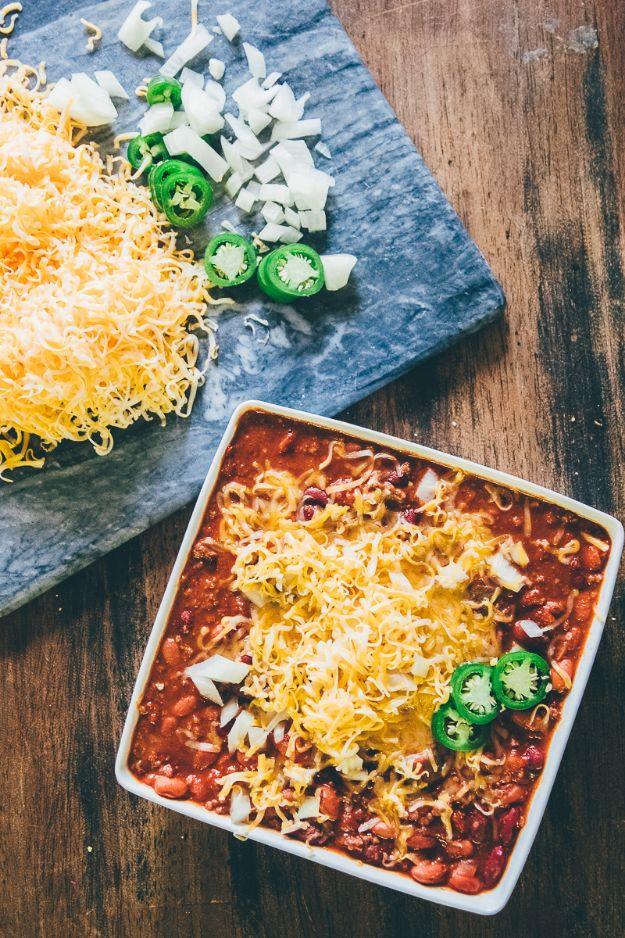 Spicy crock pot chili in a square white dish under shredded cheese, diced onion and jalapenos on slate