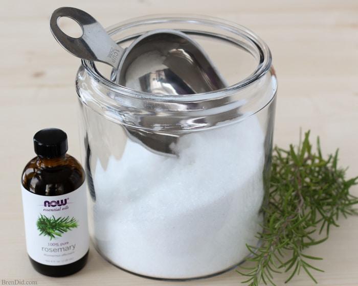 Wide glass jar of pale homemade bath salts with dried flowers and a wooden scoop resting on a folded towel