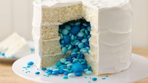 White frosted gender reveal cake cut open with blue candies pouring out of the center onto the cake board