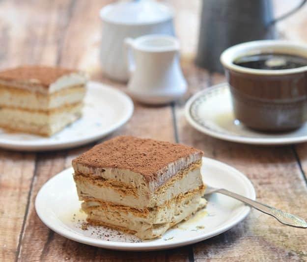 Two espresso icebox cake slices dusted with cocoa on white plates, a cup of coffee and a creamer behind
