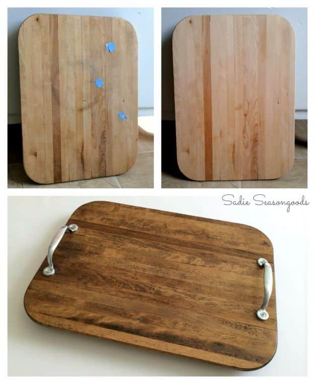 A wooden cutting board fitted with dark metal handles sits on an ottoman, styled with a candle and a plant