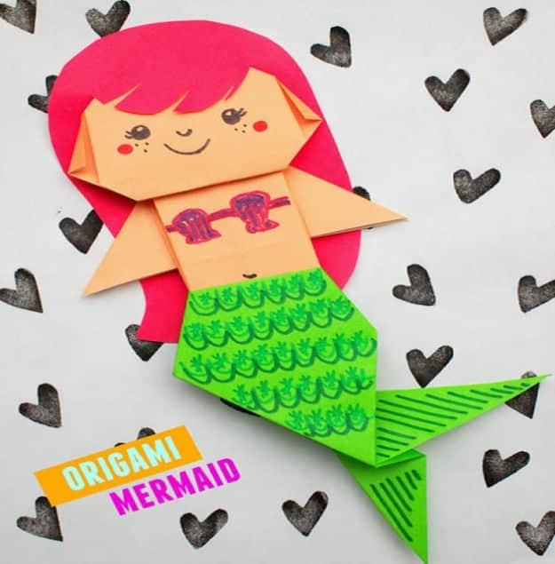Folded origami mermaid with pink hair, a peach paper face and a green scaled tail, on white paper printed with hearts