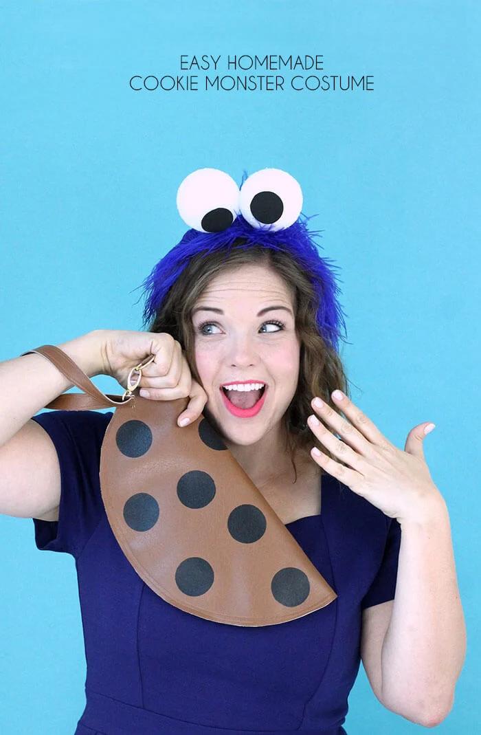Adult wearing a blue Cookie Monster eye headband and holding a brown chocolate-chip cookie clutch against a blue backdrop