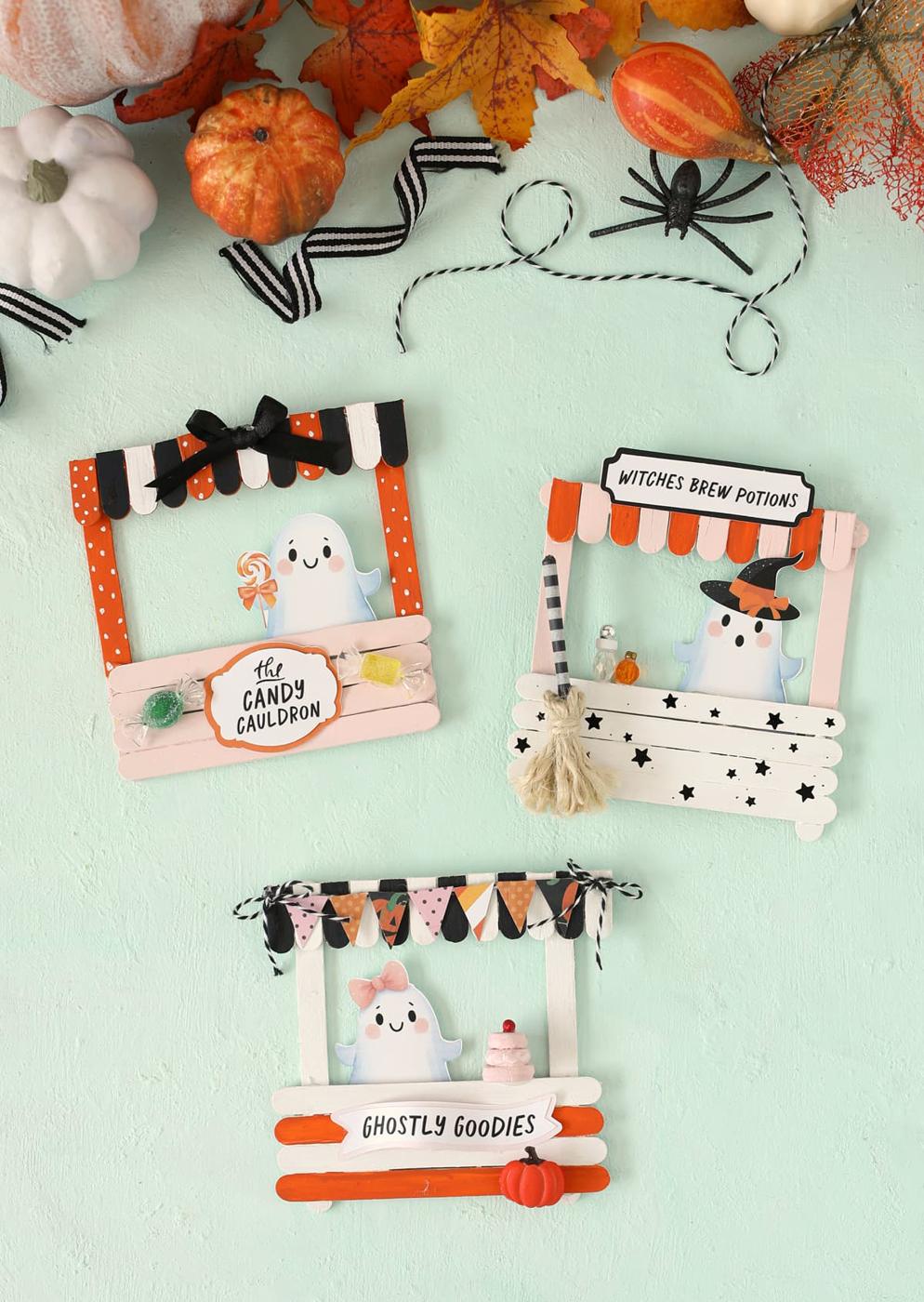 Three painted popsicle-stick Halloween storefronts with ghost characters, awnings and signs for candy, potions and goodies