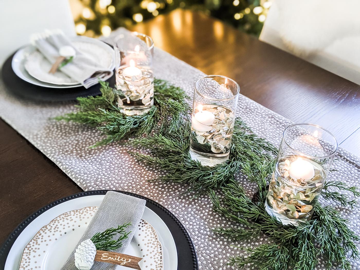 Three glass tumblers with dried eucalyptus and floating candles, each ringed by a small faux cypress wreath on a gray runner