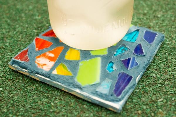 Tile mosaic coaster set with red, orange, yellow, green and blue glass pieces in grey grout, holding a glass jar on grass