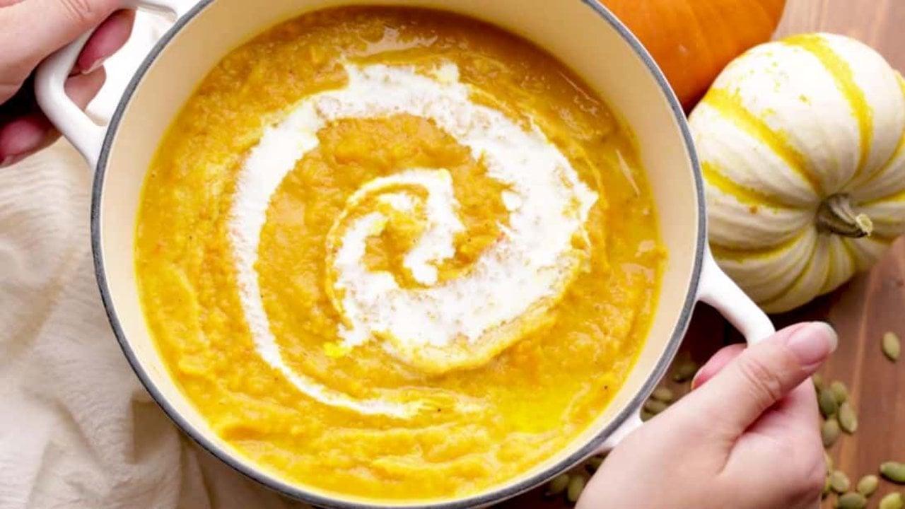 Wide pot of creamy roasted pumpkin soup with a swirl of cream, two hands holding it, squash alongside