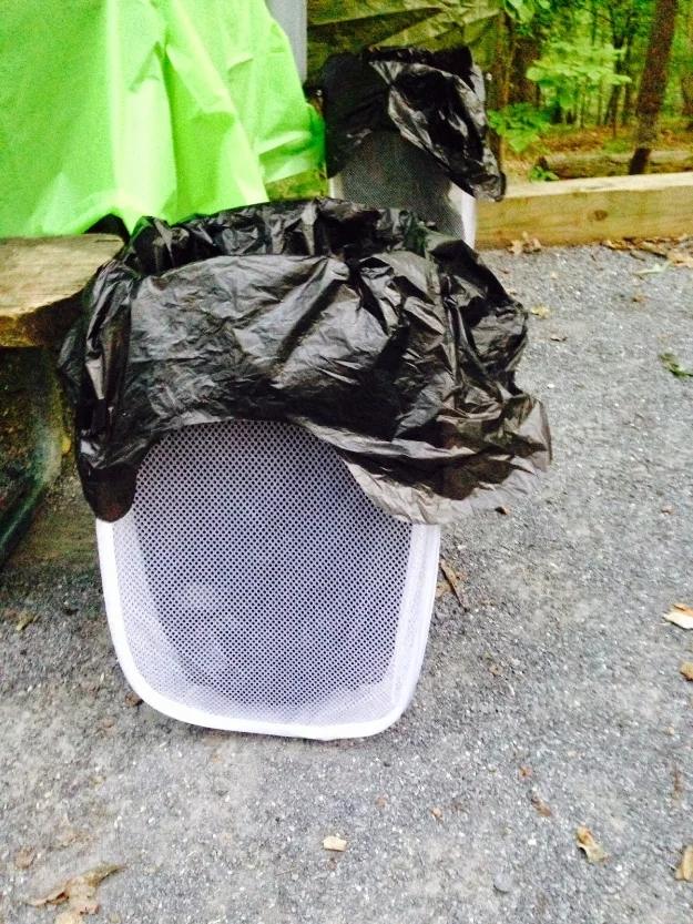 Dollar store camping trash can, a black bag pulled over a white mesh pop up hamper at a gravel campsite