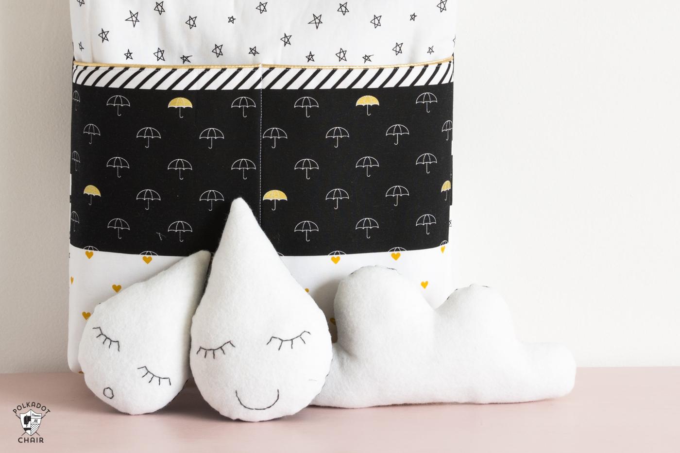 Two white fleece raindrop softies and a cloud with embroidered sleepy faces in front of a black and white umbrella print bag