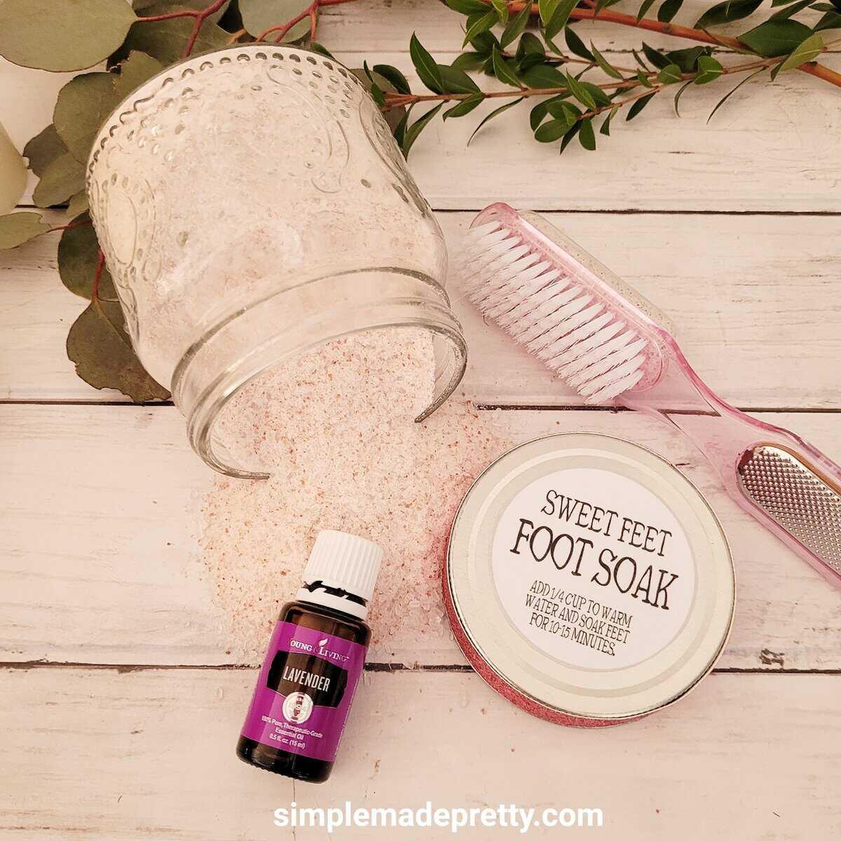 Glass jar of pink foot soak salts with a Sweet Feet label, a small brush and a bottle of essential oil on a white table