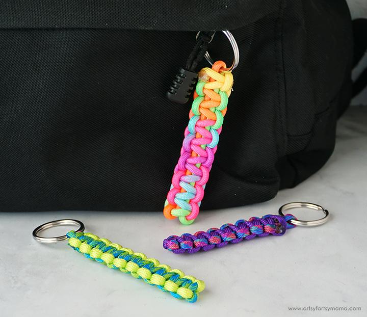 Three colorful paracord keychains in rainbow, purple and green cobra knots with silver rings beside a black backpack
