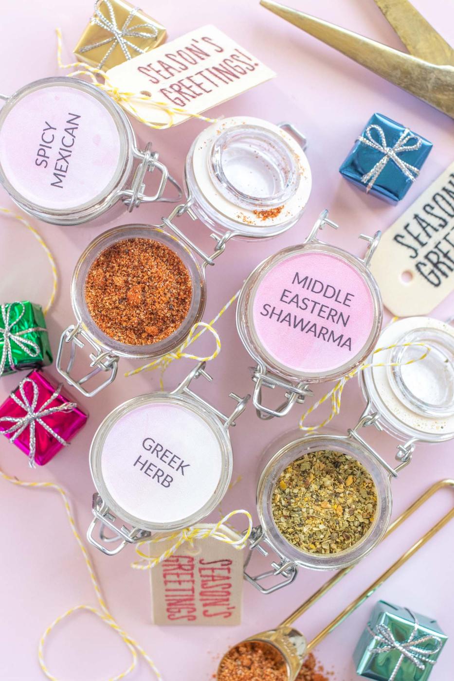 Six small hinged glass jars of red and green spice blends with printed round labels on a pink flat lay