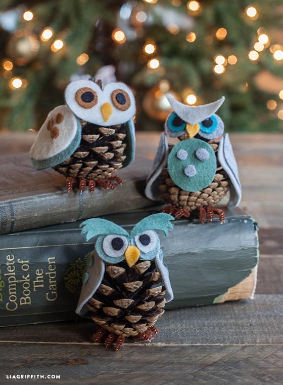 Three pine cone owls with felt wings, big round felt eyes and small beaks perched on old books in front of tree lights