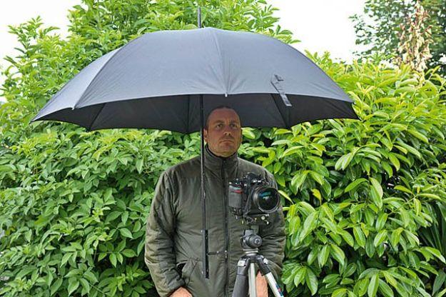 DIY umbrella holder, a man behind a tripod mounted camera with a large gray umbrella fixed above it in front of a green hedge