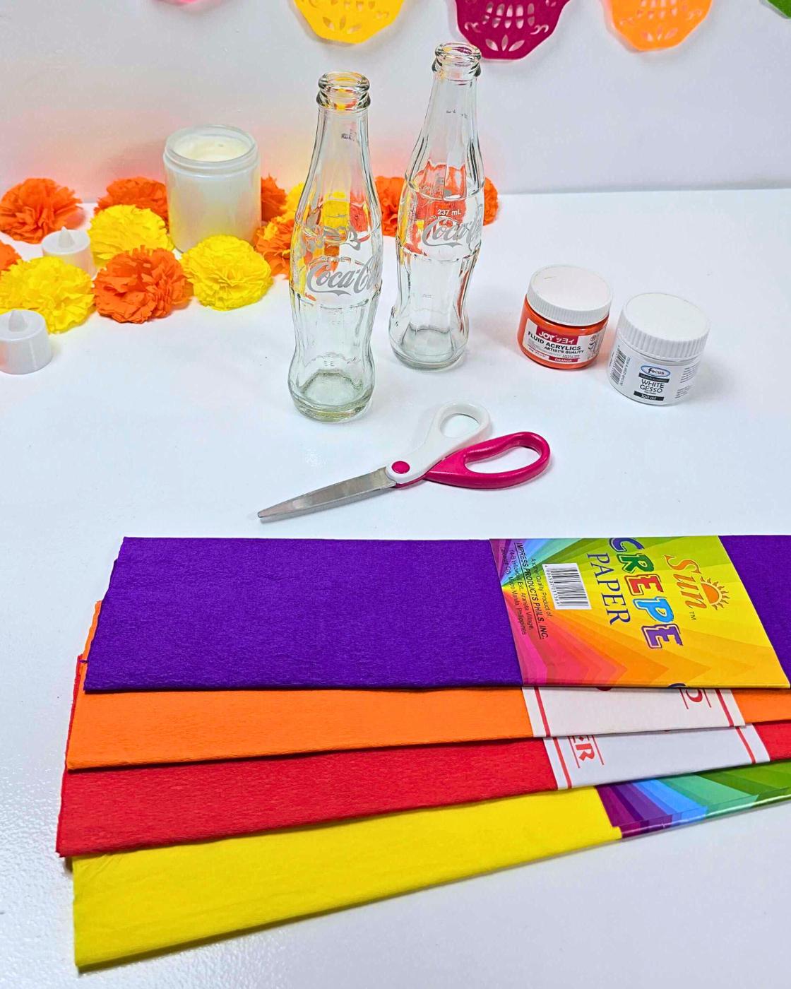 Easy Crafts - supplies laid out for the project, two empty glass Coca-Cola bottles, a jar of orange paint, a jar of white primer, pink-handled scissors and four rolled crepe paper 