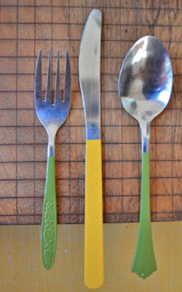 A fork, a knife and a spoon with handles spray painted moss green and mustard yellow on a wooden board