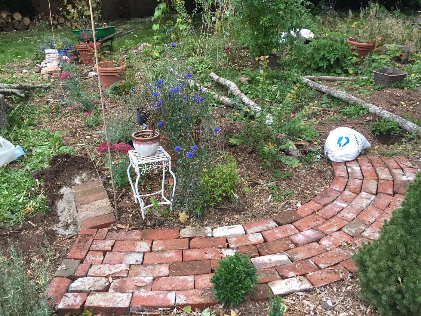 Curving red brick path laid in sand, winding through a planted garden bed toward a distant flowering border