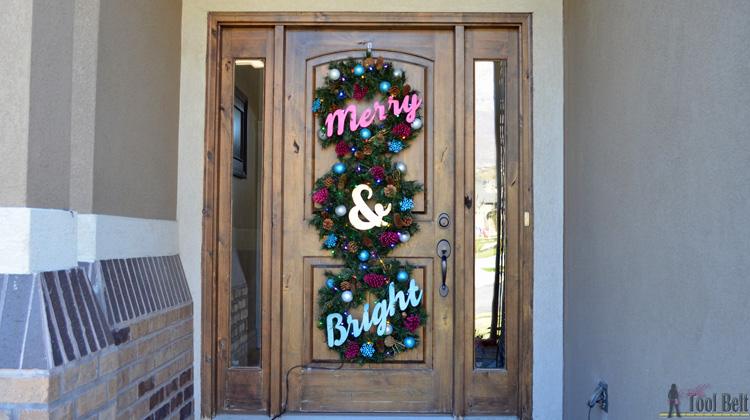 Three evergreen wreaths stacked down a tall wood front door with plywood letters reading Merry and Bright in pink and teal
