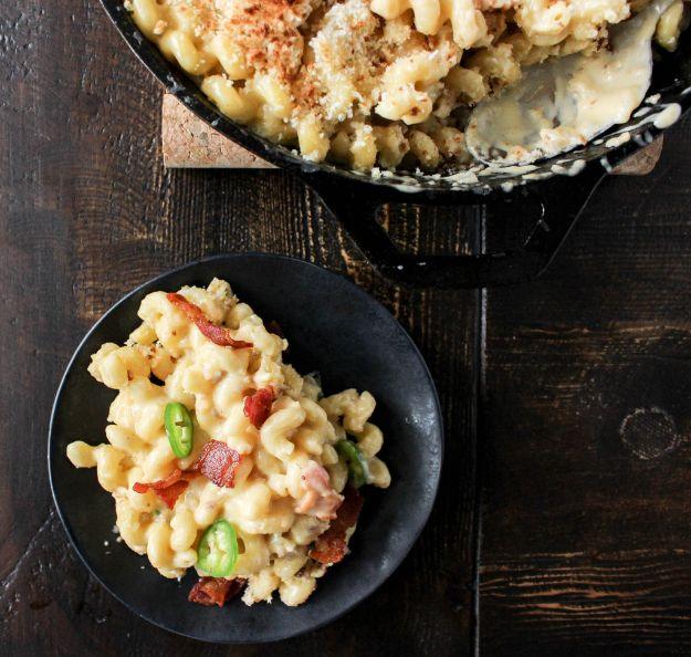 Jalapeno bacon mac and cheddar served on a black plate with the cast iron skillet behind it and sliced peppers