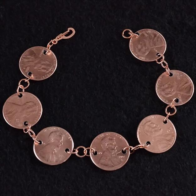 Penny bracelet of seven drilled copper coins linked with jump rings and a clasp, laid flat on black fabric
