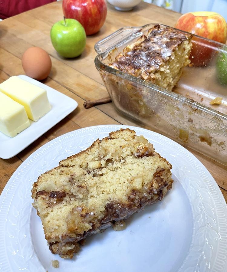 Breakfast Bread Recipes - single slice of cinnamon apple bread on a white plate with the rest of the loaf in its glass dish, butter, an egg and apples behind
