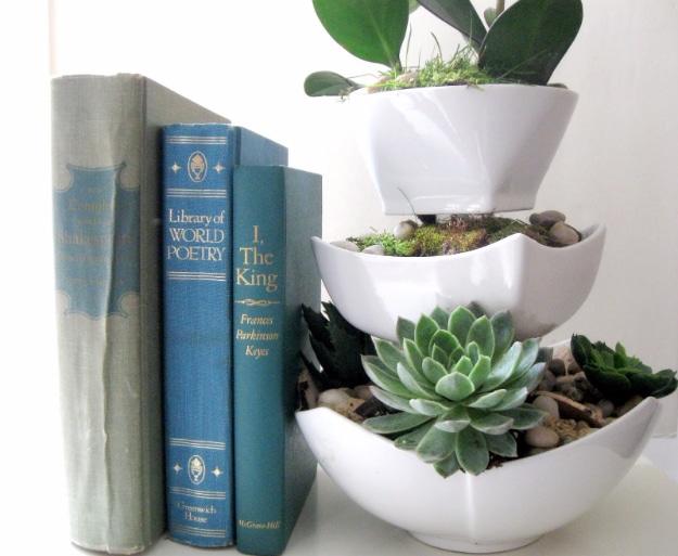 Three tier planter of stacked white ceramic bowls and clear glasses planted with succulents, moss and river rocks