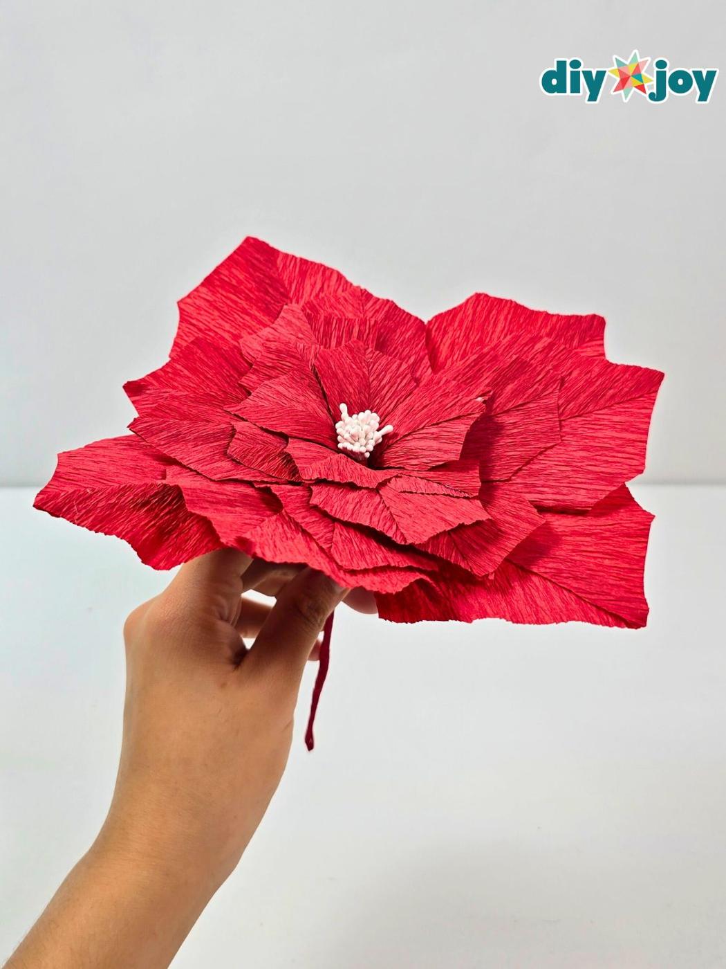 Hand holding a large red crepe paper poinsettia with layered petals and a pale stamen center against a white wall
