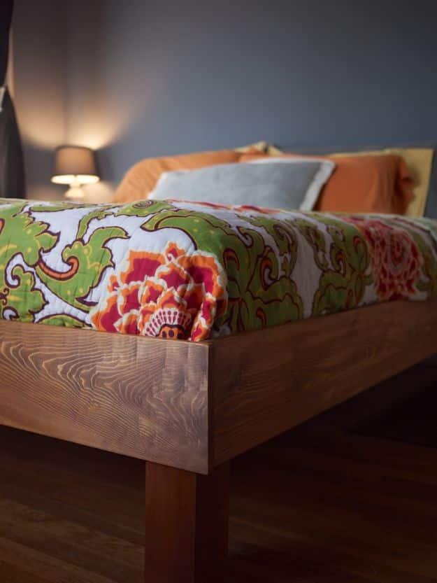Corner of a stained wood king bed frame with a thick side rail, square leg and green and red floral bedding
