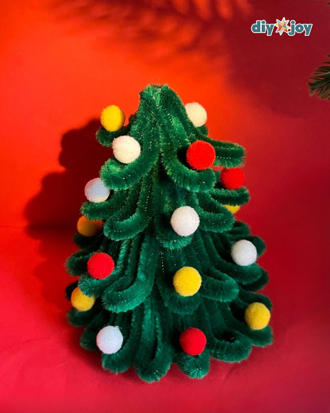 Pipe Cleaner Christmas Tree - one finished green tree covered in red, yellow, and white pom poms casting a shadow on a solid red background