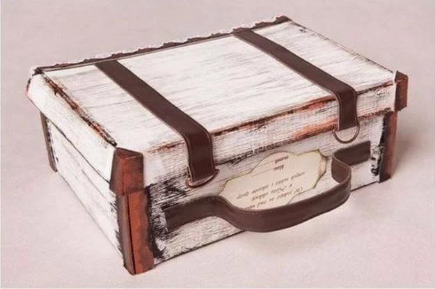 Shoe box painted distressed white with brown paper straps, corner protectors, a handle and a luggage tag