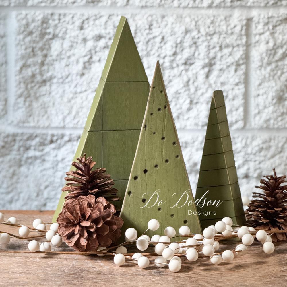 Three olive green scrap wood triangle trees with grid lines, drilled dots and stripes beside pinecones and white berries