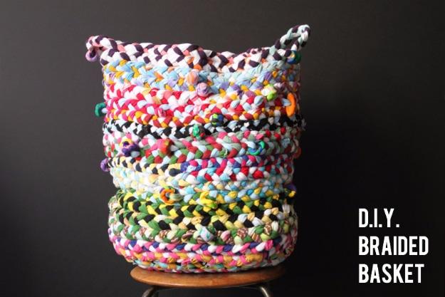 Tall coiled basket braided from multicolored t-shirt strips with two small handles, standing on a stool against a dark wall