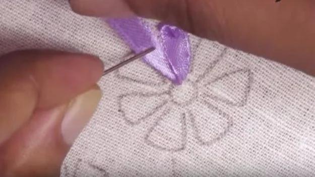 Close up of a hand guiding a needle and purple silk ribbon through linen along a pencilled flower petal outline